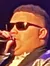 A performer wearing sunglasses and a shiny jacket sings into a microphone on stage, with a vibrant, colorful light display in the background.