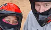 Two people wearing helmets are posing for a selfie in front of parked ATVs on a dirt surface.
