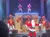 A festive stage show features a person dressed as Santa Claus, singers, and dancers with holiday decorations.