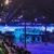 The image shows an indoor stage set with a winter theme, featuring snowy landscapes and log cabins, surrounded by an audience seating area.
