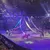 The image shows a vibrant indoor equestrian performance with riders on horses weaving through hanging ribbons, surrounded by an audience in a theater setting.