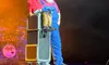 A ventriloquist, dressed in overalls, is performing on stage with a cowboy puppet, under colorful stage lights.