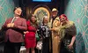 Five people are posing happily in front of a dartboard while dressed in a mix of formal and themed attire, beneath a sign that says 