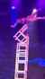 A performer balances precariously atop a stack of chairs under dramatic lighting on stage.
