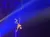 A performer is suspended upside down playing a guitar above an audience at an indoor event with colorful lighting.