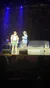 Two performers are on stage under blue spotlights, with one dressed in lederhosen and red suspenders and the other in a blue jacket and cowboy hat, speaking in front of an audience.