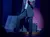 A performer wearing a black cowboy hat and a jacket with red roses is singing on stage under blue lighting.