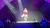 A performer in a colorful dress sings on stage under bright spotlights, surrounded by a band against a starry backdrop.