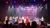 Performers dressed in patriotic costumes stand on stage in front of a large American flag, while an audience watches.