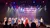 Performers in red, white, and blue costumes stand on stage with an American flag backdrop, while an audience watches.