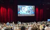 A large audience is seated in a theater with a sign reading 