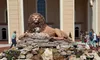 A large lion and lamb sculpture is displayed on a rocky fountain in front of a beige building, surrounded by flowers and visitors.