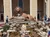 A large lion and lamb sculpture is displayed on a rocky fountain in front of a beige building, surrounded by flowers and visitors.
