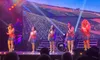 Five women in matching sparkly dresses are performing on stage with colorful lighting and a drum set in the background.