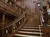 The image depicts an ornate, wooden staircase featuring intricate carvings, gold detailing, and a decorative statue holding a lamp, all under a domed skylight.