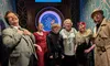 A group of six people wearing a mix of vintage and casual attire pose cheerfully in front of a dartboard in a venue decorated with geometric patterns.