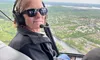 A person wearing sunglasses and a headset is sitting in a helicopter, flying over a scenic landscape with a river and greenery below.