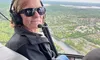 A person wearing sunglasses and a headset is flying in a helicopter, with a scenic view of the landscape below.