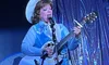 A person in a blue and white western outfit is performing on stage, playing a guitar and singing into a microphone.