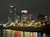 The image shows a nighttime cityscape with illuminated skyscrapers and waterfront buildings reflecting on the water.