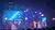 The image shows a live musical performance with musicians dressed in white, playing string instruments on a stage illuminated by blue and white lights, with digital graphics displayed on a large screen in the background.