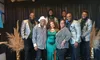 A group of people, dressed in stylish attire with matching silver jackets, smile together on a decorative stage.