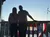 Two silhouetted people stand on a balcony with a red, white, and blue banner and a sunset sky in the background.