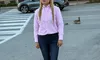A person in a pink shirt and jeans stands on a street with a building and parked car in the background, and geese nearby.