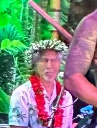 A performer in traditional attire is energetically dancing with a stick, while a person beside him appears to be singing or speaking into a microphone, set against a vibrant, tropical-themed backdrop.