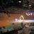 A live action performance is taking place in an arena, featuring a ring of fire and a blurred figure moving rapidly, with an audience watching from the stands.