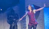 A performer dressed in a red plaid outfit energetically dances on stage near a statue of a knight.