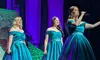Three women in elegant teal gowns perform on stage with a microphone, surrounded by colorful lighting and mist.