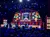 The image shows a live performance at the Grand Ole Opry stage, featuring musicians playing under vibrant lighting and a decorative backdrop.