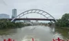 A red paddlewheel boat is moving down a river towards a modern arched bridge, with city buildings in the background.