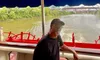 A man wearing a cap and glasses leans on the railing of a riverboat with red paddle-wheels, cruising along a river with a bridge and greenery in the background.