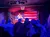 A performer is singing on stage in front of an American flag backdrop, with an audience seated and watching in dim lighting.