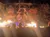 A performer stands on stage amidst a pirate-themed set, with flames dramatically rising in front of an audience.
