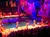 The image shows a vibrant stage performance with performers dressed in colorful costumes, set against a backdrop of a pirate-themed stage with a ship and an audience watching from tiered seating.