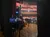 The image shows a backstage view of an empty auditorium with red seats, stage equipment, and a sign indicating 