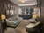 The image shows an elegant dressing room with a seating area, framed photos on the wall, a large mirror surrounded by lights, and a cozy, carpeted floor.