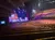 The image shows an empty theater with wooden pews and a stage lit up with purple and blue lights, featuring the words 