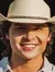 A person wearing a cowboy hat stands smiling with a guitar case in front of the 