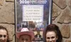 Three people, two in matching patriotic costumes and one in a yellow outfit with a pink hat, pose together in front of a stone wall with a concert poster.