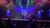 A singer performs on stage with colorful lighting and projected angel wings in the background, accompanied by three musicians sitting around him.