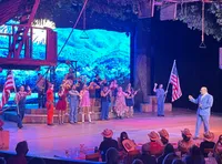 A lively theatrical performance is taking place on stage, featuring performers in colorful costumes and an elaborate set resembling a treehouse, with an audience watching from their seats.