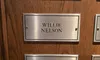 The image shows a wooden wall with metal plaques engraved with names of notable individuals such as Willie Nelson, Dolly Parton, and others.