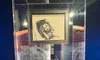 The image shows a display case containing a drawing and a necklace labeled as the 