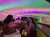 People are seated on a boat ride under a tunnel illuminated with vibrant purple and green lights.