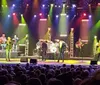 Four men are performing on stage with microphones, one of them wearing a cowboy hat, against a backdrop of drums and stage lights.