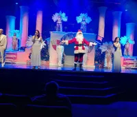 A person dressed as Santa Claus is waving on stage against a blue backdrop.